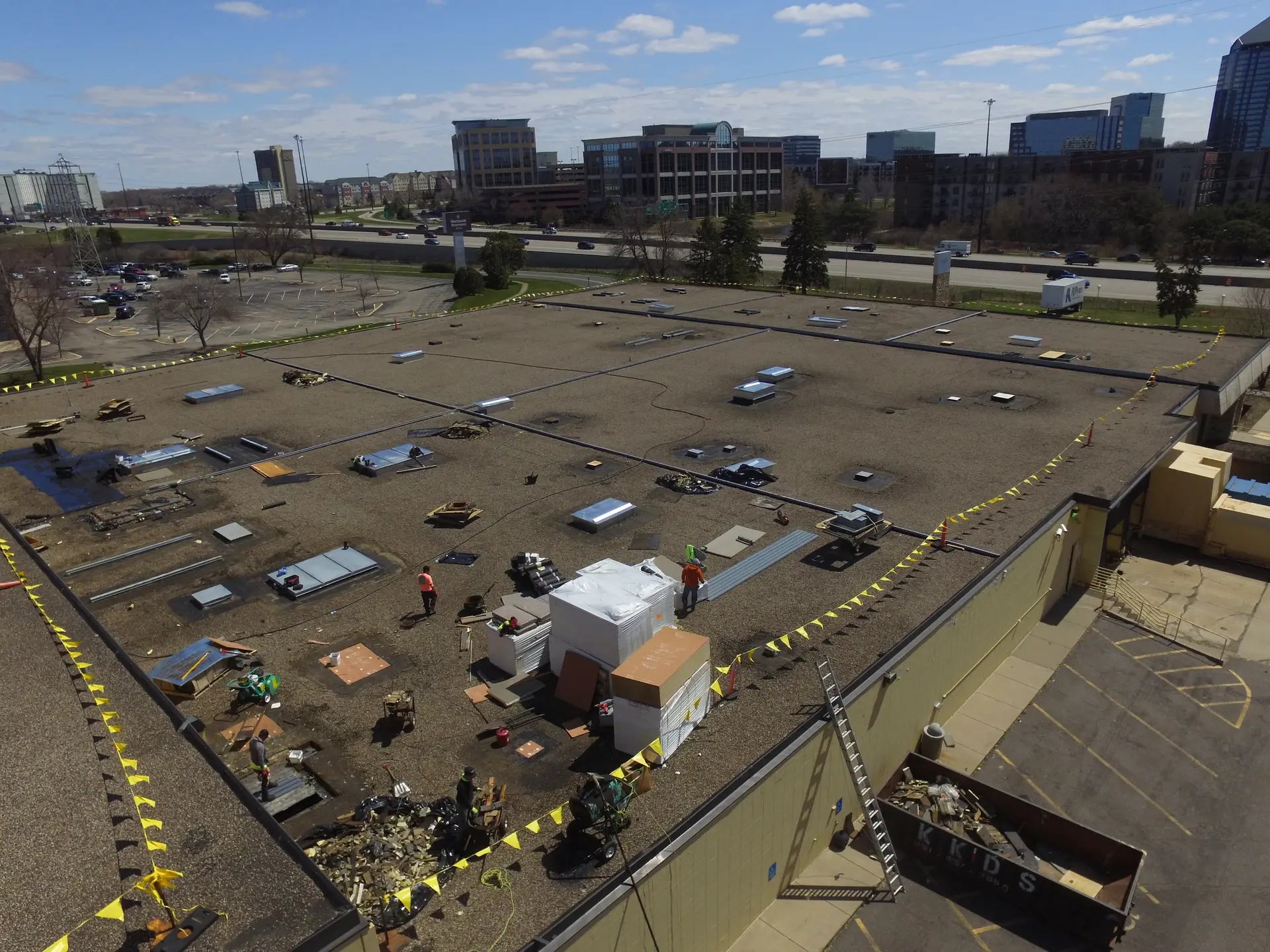 Crew working across a large flat commercial roof, materials staged inside caution-taped zones