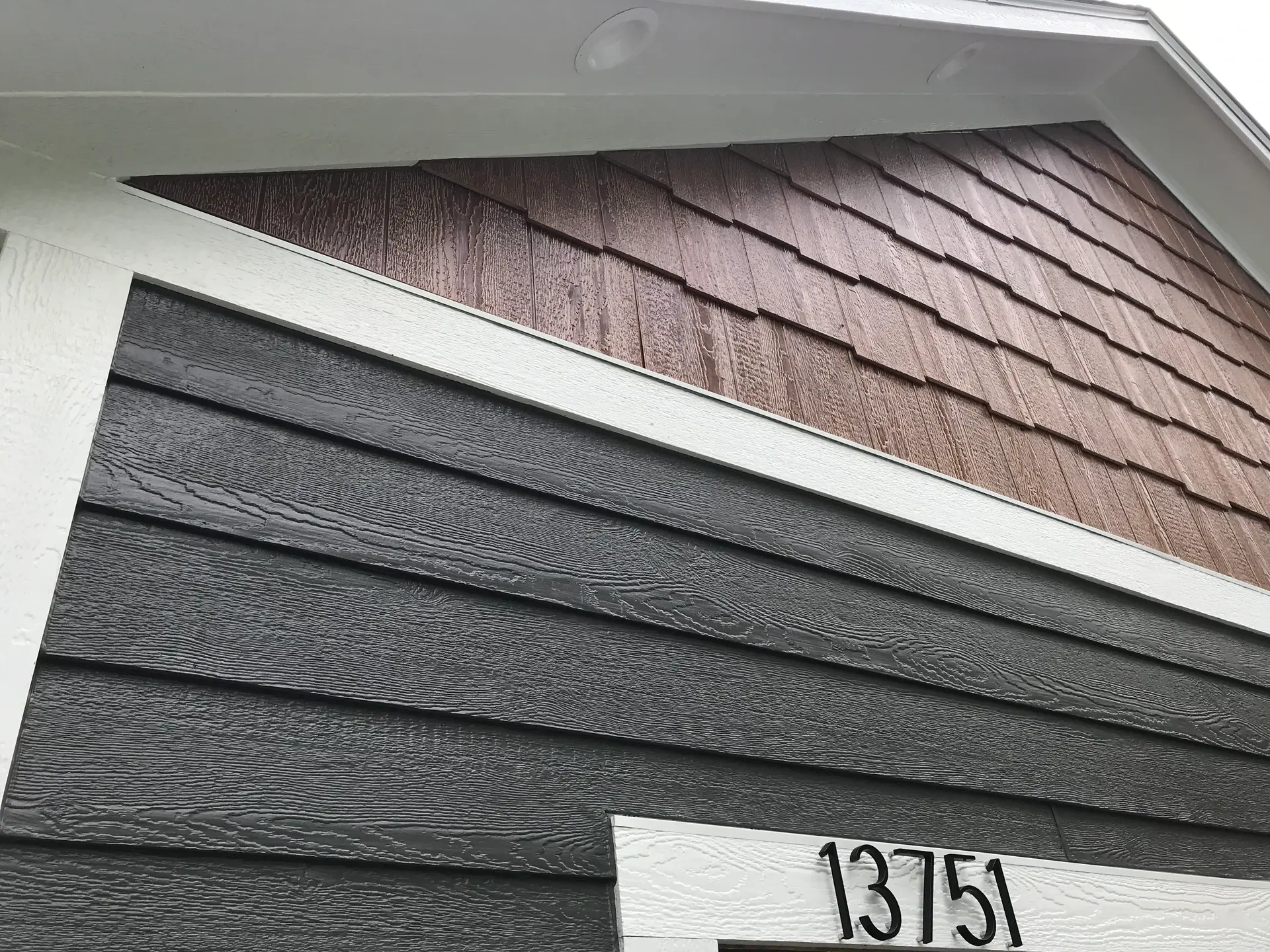 Dark gray lap siding meeting a shake-style gable above a house number