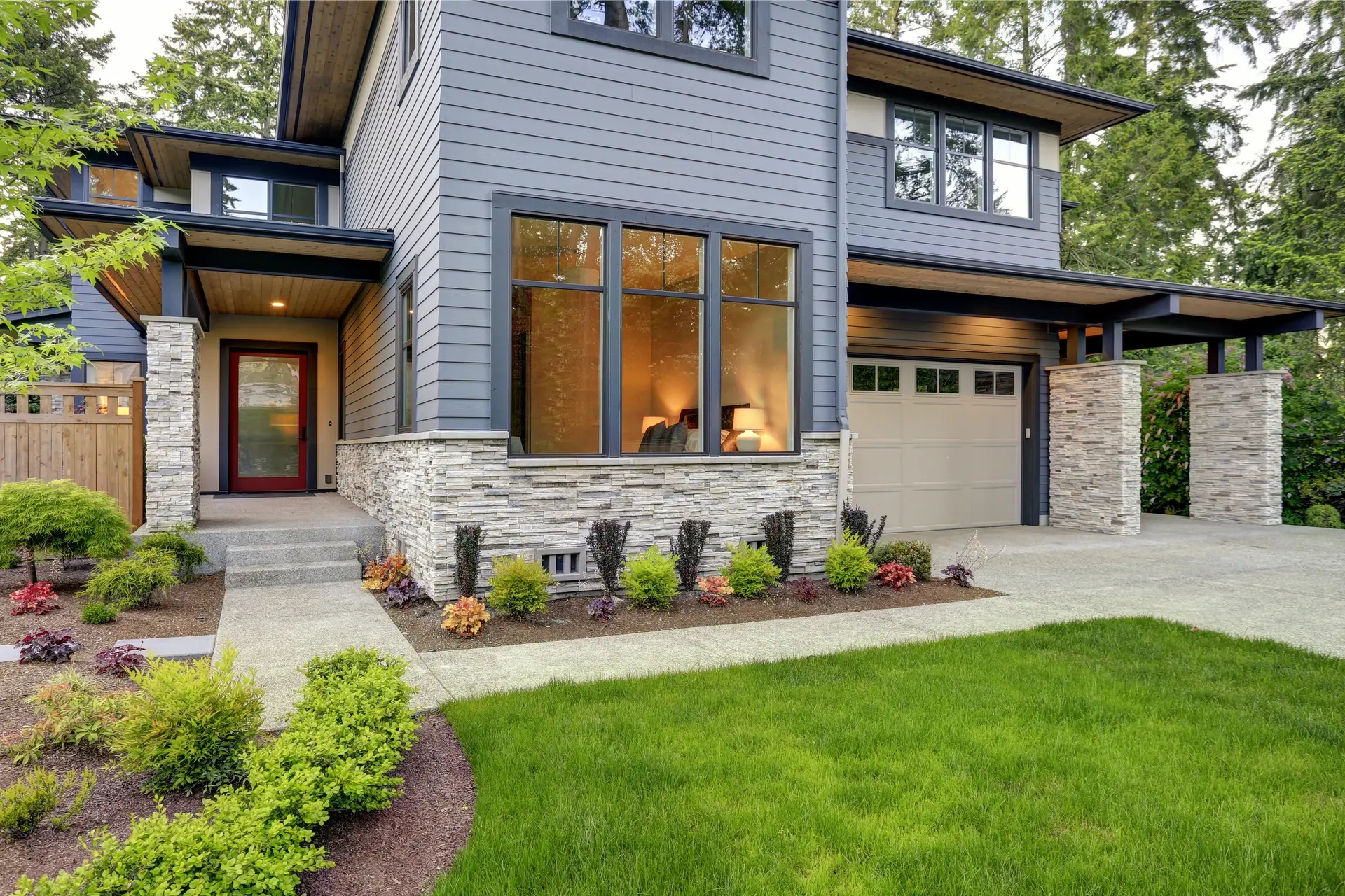 Large windows lit from inside a two-story home with gray siding and stone veneer